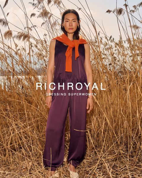Une femme en chemise bleu clair et pantalon gris tient une boule à neige devant un œil. Elle pose dans une pièce aux murs en bois avec une affiche de ski et porte de grandes boucles d'oreilles. Le texte est "RICHROYAL DRESSING SUPERWOMEN".