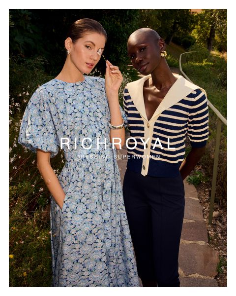 Une femme en chemise bleu clair et pantalon gris tient une boule à neige devant un œil. Elle pose dans une pièce aux murs en bois avec une affiche de ski et porte de grandes boucles d'oreilles. Le texte est "RICHROYAL DRESSING SUPERWOMEN".