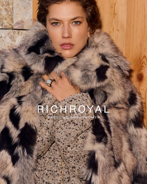 Une femme en chemise bleu clair et pantalon gris tient une boule à neige devant un œil. Elle pose dans une pièce aux murs en bois avec une affiche de ski et porte de grandes boucles d'oreilles. Le texte est "RICHROYAL DRESSING SUPERWOMEN".