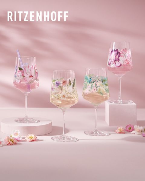 Un ensemble de verres Ritzenhoff colorés et artistiques, remplis de boissons. Chaque verre a un design unique avec des motifs et des figures vibrantes. L'arrière-plan présente des reflets de lumière et d'autres verres, créant une atmosphère festive.