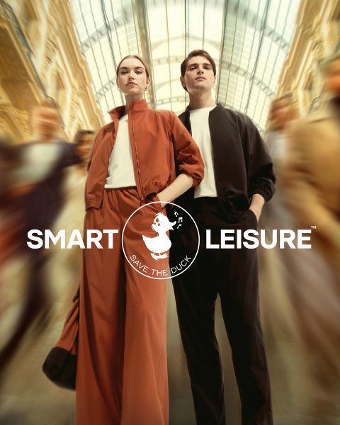 Un mannequin défile sur un chemin couvert de feuilles. Elle porte une veste de fourrure orange, une jupe assortie et un chapeau avec des collants orange et des chaussettes couleur pêche. Le logo de « Save the Duck » est visible dans le coin.