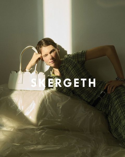 Personne portant un blazer noir sur une chemise rayée et un jean bleu, tenant un sac à main noir. Le texte "JULIA SKERGETH" est clairement visible superposé à l'image.