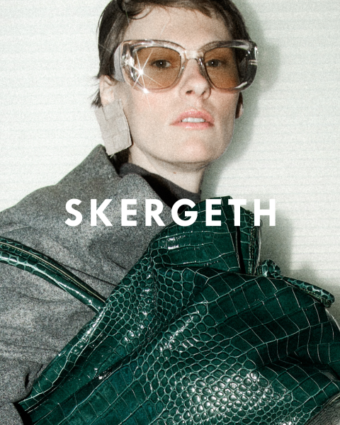 Personne portant un blazer noir sur une chemise rayée et un jean bleu, tenant un sac à main noir. Le texte "JULIA SKERGETH" est clairement visible superposé à l'image.