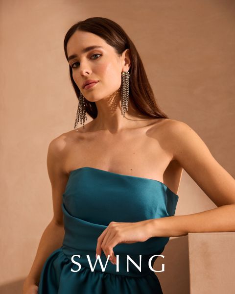 Une femme vêtue d'une robe de soie vert sauge est assise sur une chaise couleur crème devant un fond floral. Elle porte des talons dorés, des boucles d'oreilles voyantes et un bracelet. Le mot "SWING" apparaît en bas de l'image.