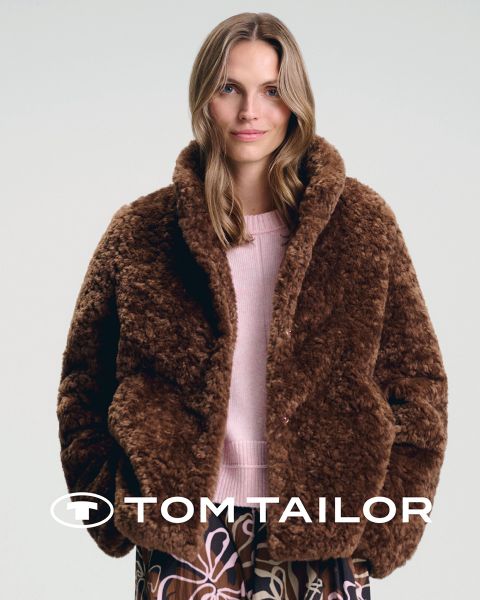 Une femme souriante portant un pull marron et un homme portant un pull bleu marine se tiennent côte à côte sur une plage. Ils portent tous les deux des jeans. Le ciel est clair et bleu. Le logo "Tom Tailor" apparaît en bas.