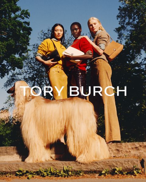 Une femme portant un haut scintillant et une jupe verte est assise à côté de boîtes cadeaux vertes et tient un sac Tory Burch à fleurs à la main ; derrière elle se trouve un arbre de Noël décoré.