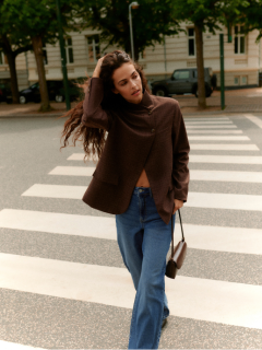 Une femme aux cheveux bouclés se tient devant un mur carrelé. Elle porte un cardigan gris clair duveteux, un short en cuir noir taille haute et des collants noirs, les mains dans les poches et une expression confiante sur le visage.