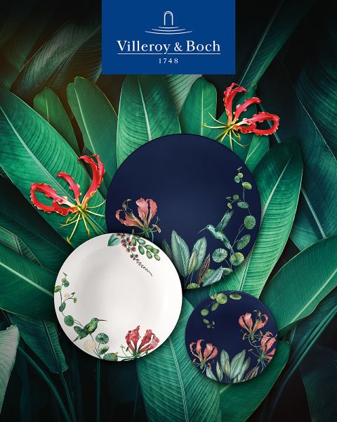 Sur une table basse en verre réfléchissant, on trouve une tasse de café dans une tasse blanche et dorée, une petite théière, un pot à lait, une bougie et une assiette de biscuits. La scène est élégante et confortable, et le logo de Villeroy & Boch est visible en bas.