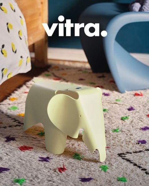 Un salon moderne comprend un canapé rayé rose et beige, une chaise blanche avec des pieds en bois et une table basse ronde en bois. Sur la table, il y a un vase avec des fleurs colorées, au-dessus il y a « vitra. » au mur. Le sol est recouvert d'un tapis doux.