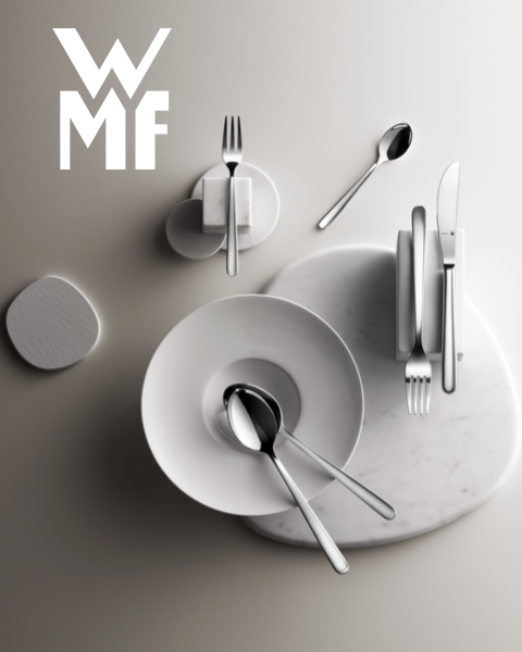 Une cocotte remplie de figues, de mûres et de garnitures aromatiques est posée sur une planche de bois sombre. Au-dessus de la cocotte, on aperçoit le logo WMF. L'arrière-plan est une surface sombre texturée.