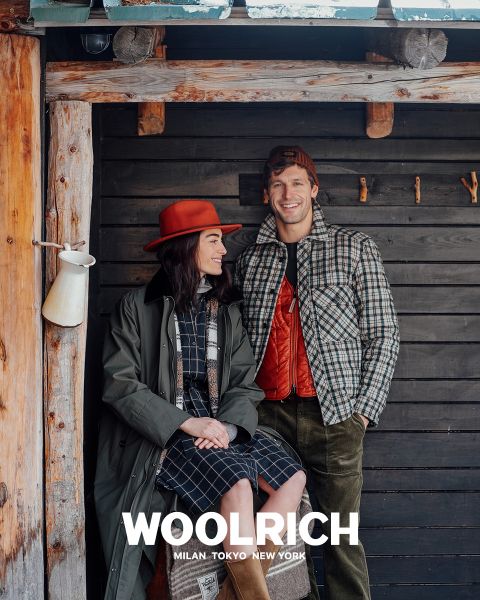 Une personne portant un manteau d'hiver marron clair, un bonnet bleu et une chemise à carreaux bleus sur un sweat à capuche se tient devant un fond uni. En dessous apparaît le mot « Woolrich » ainsi que les villes de Milan, Tokyo et New York.
