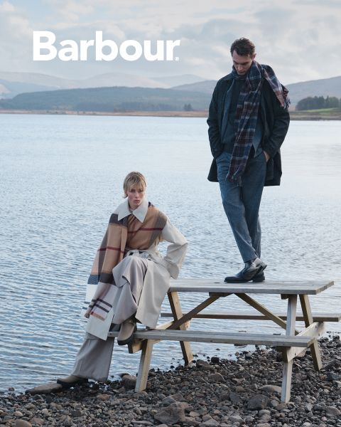 Deux personnes en vêtements Barbour élégants et multicouches et écharpes à carreaux posent au bord d'un lac. L'une est assise sur une table de pique-nique, tandis que l'autre est debout, avec des montagnes et de l'eau en arrière-plan. Le logo Barbour apparaît en haut à gauche.