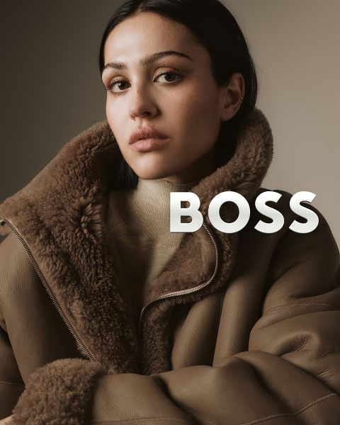 Une femme portant un manteau en peau lainée marron regarde l'appareil photo avec assurance devant un fond neutre. Le mot "BOSS" est affiché en lettres blanches grasses à côté de son épaule.