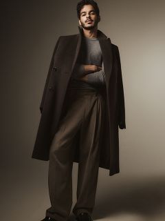 Un homme se tient là avec assurance, portant un long manteau sombre sur un pull gris et un pantalon marron à jambes larges, les bras croisés. L'arrière-plan est simple et doucement éclairé, créant une atmosphère élégante et sophistiquée.