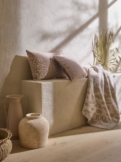 Un espace minimaliste présente un banc en pierre beige orné de deux coussins texturés bruns et beiges, d'un jeté à motifs et d'herbes séchées. Un panier tressé, un vase en céramique rustique et un grand pichet décorent le sol sous une douce lumière naturelle.
