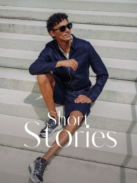 Un homme portant des lunettes de soleil, une veste bleu marine et un short est assis en souriant sur un escalier extérieur. Les mots "Short. Stories" apparaissent en bas.