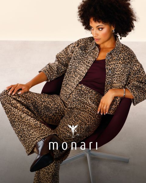 Une femme souriante portant un manteau beige, une chemise rayée, un jean bleu et une écharpe claire se tient à l'extérieur devant un grand bâtiment. Le logo Monari est visible en bas de l'image.