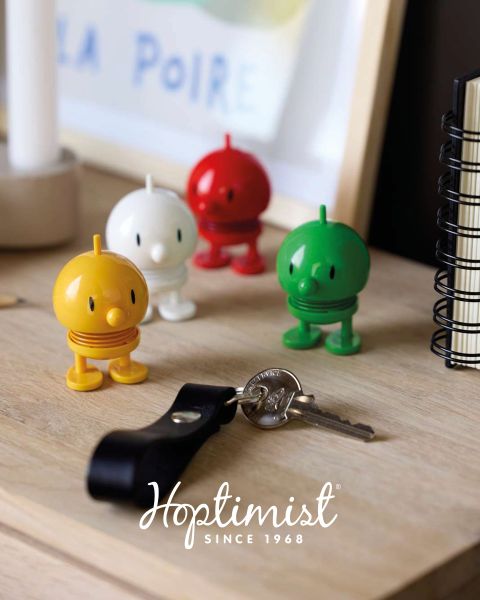 Quatre figurines Hoptimist colorées sur un bureau en bois à côté d'un carnet, d'une lampe et d'un trousseau de clés avec le texte "Hoptimist Since 1968" en bas.