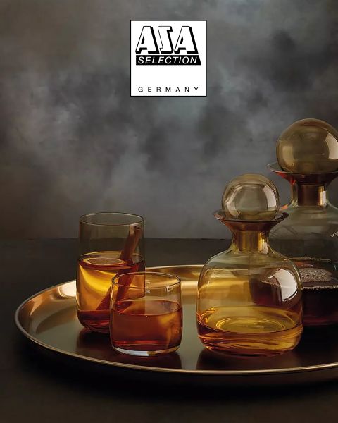 Des carafes couleur ambre avec des verres assortis, remplies d'un liquide brun, sont posées sur un plateau doré devant un fond texturé gris et atmosphérique. En haut, le logo d'ASA Selection Germany est représenté.