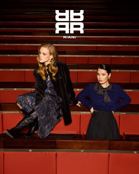 Deux femmes posent avec style dans un théâtre aux sièges rouges. L'une est assise sur le dossier d'un siège et porte une robe à motifs, un manteau noir et des bottes. L'autre se tient à côté d'elle en pull bleu marine et jupe noire. Au-dessus d'elles apparaît le logo "RIANI".