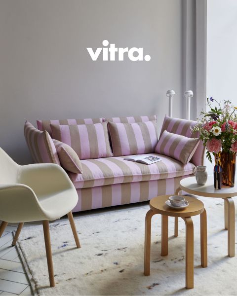 Un salon moderne comprend un canapé rayé rose et beige, une chaise blanche avec des pieds en bois et une table basse ronde en bois. Sur la table, il y a un vase avec des fleurs colorées, au-dessus il y a « vitra. » au mur. Le sol est recouvert d'un tapis doux.