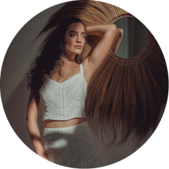 Une femme vêtue d'un haut sans manches blanc et d'une jupe se tient au soleil, un bras posé sur sa tête, à côté d'un miroir mural rond à franges, derrière lequel des ombres se projettent.