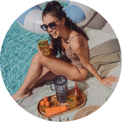 Une femme en maillot de bain à motif léopard est assise sur une serviette au bord de la piscine, souriante et tenant une boisson à la main. Elle porte des lunettes de soleil et se trouve à côté d'un plateau avec des boissons, de la crème solaire et des fruits. Derrière elle, on aperçoit une bouée gonflable dans l'eau.