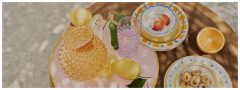 Une table en bois avec une cruche en verre texturé, un verre rose, des citrons entiers avec des feuilles, des assiettes décoratives empilées, une demi-orange et une assiette de petits biscuits ronds dans la lumière du soleil tachetée.
