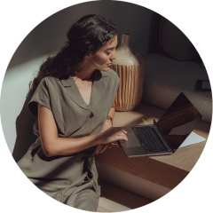 Une femme aux longs cheveux noirs est assise par terre et utilise un ordinateur portable posé sur une petite table en bois. Elle porte une robe kaki à manches courtes, éclairée par la lumière du soleil venant de la gauche. Derrière elle, on aperçoit un grand vase et un canapé.