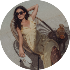 Une femme en robe jaune clair et portant des lunettes de soleil se détend à l'extérieur sur une chaise ronde en osier. Elle tient une pochette blanche à la main et est assise à côté d'une table avec un pichet en verre, des tasses et une orange. La lumière du soleil projette des ombres sur la scène.