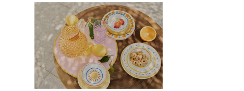 Une table ronde en bois avec des assiettes colorées, un bol de biscuits, des oranges coupées en deux, des citrons et de la verrerie texturée, le tout disposé sur une nappe à rayures roses dans la lumière naturelle du soleil.