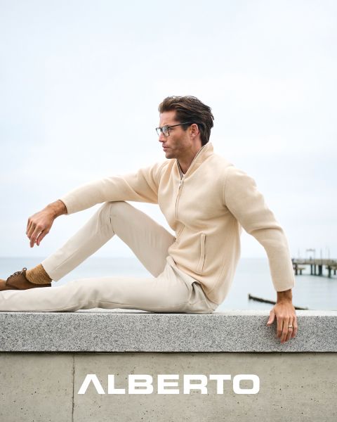 Un homme portant des lunettes, un pull beige zippé et un pantalon clair est assis sur un promontoire rocheux au bord de la mer. Il regarde sur le côté, et en dessous de lui, on peut lire "ALBERTO" en lettres blanches. L'arrière-plan est nuageux et on aperçoit une jetée.