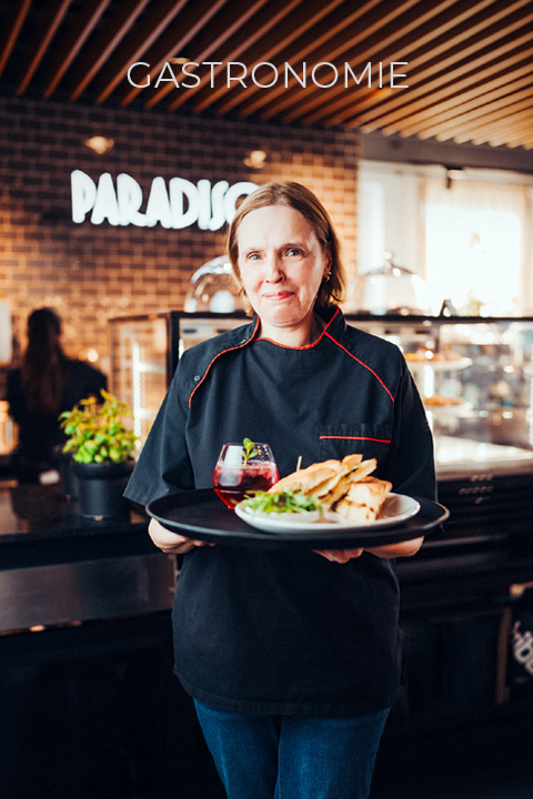 Un cuisinier en uniforme noir tient un plateau avec un plat et une boisson à la main. Il se tient dans un restaurant moderne où l'on aperçoit les panneaux "PARADISO" et "GASTRONOMIE" en arrière-plan.