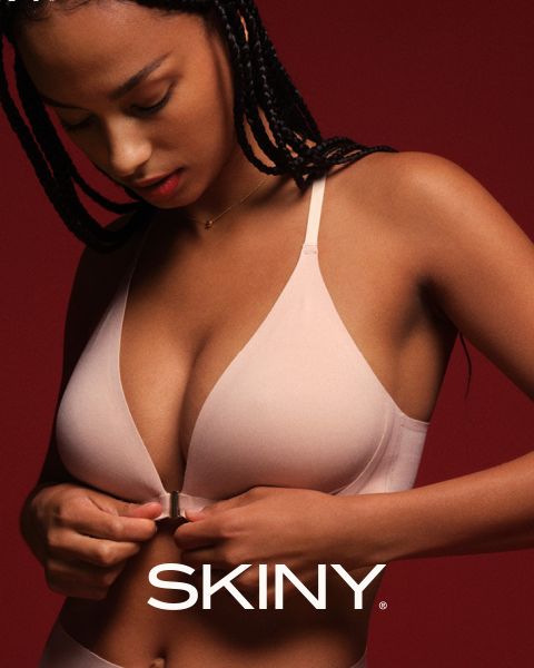 Une femme aux cheveux tressés ferme un soutien-gorge rose clair à fermeture frontale devant un fond rouge foncé. Le mot « SKINY » apparaît en bas de l'image.