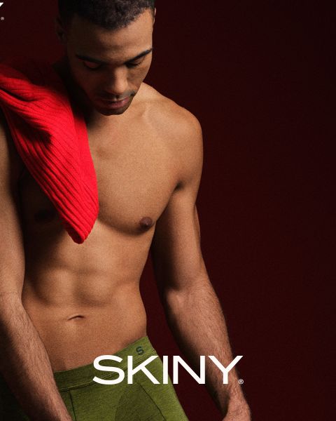 Un homme musclé et décontracté regarde vers le bas. Il porte un pull rouge sur l'épaule et a des sous-vêtements verts. Le fond est rouge foncé et le mot "SKINY" apparaît en bas.