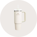 Ein cremefarbener Stanley-Becher mit Henkel, Edelstahlrand und einem Deckel mit Strohhalm, dargestellt auf einem hellgrauen, runden Hintergrund.