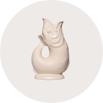 Ein glänzender, cremefarbener Keramikkrug in Form eines Fisches mit offenem Maul, strukturierten Schuppen und einem gebogenen Schwanz vor einem hellgrauen, runden Hintergrund.