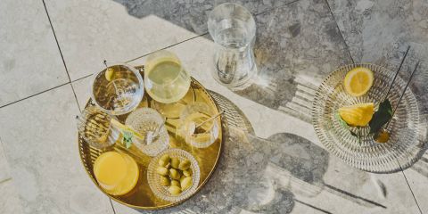 Un plateau doré avec diverses boissons, des olives et un verre de jus d'orange est posé sur une surface carrelée au soleil. À proximité, une assiette en verre contient des tranches de citron et une feuille, à côté d'une carafe en verre remplie d'eau.