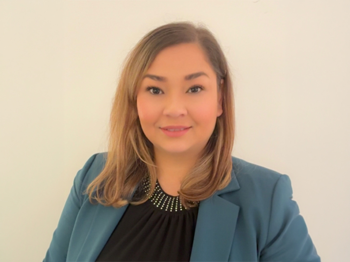 Une femme aux cheveux châtain clair mi-longs, vêtue d'un blazer bleu sarcelle et d'un haut noir, sourit devant un fond clair uni.