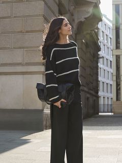 Une femme aux cheveux longs se tient sur un trottoir en ville. Elle porte un pull noir à rayures blanches et un pantalon noir à jambes larges, et tient une grande pochette noire à la main, tandis qu'elle est éclairée par le soleil venant de derrière.