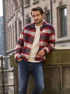 Un homme aux cheveux courts et foncés et à la barbe s'appuie sur une balustrade en pierre à l'extérieur. Il porte une veste à carreaux rouge et blanc sur un pull crème et un jean bleu. Des bâtiments urbains sont visibles en arrière-plan.