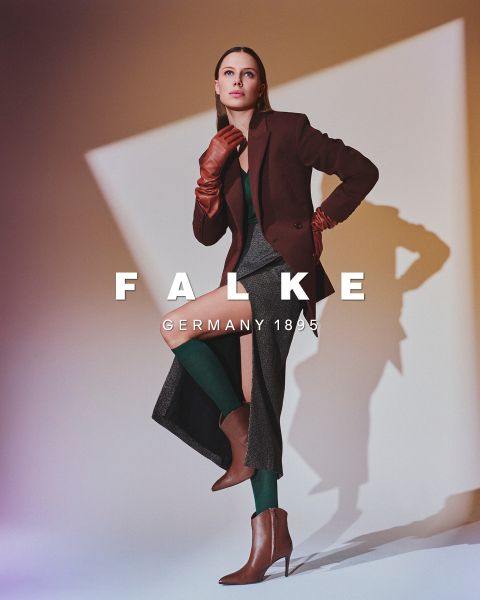 Un mannequin portant un blazer marron, des gants marron, des collants verts, une jupe grise à fentes hautes et des bottines marron pose devant un fond géométrique. Le texte indique "FALKE ALLEMAGNE 1895".