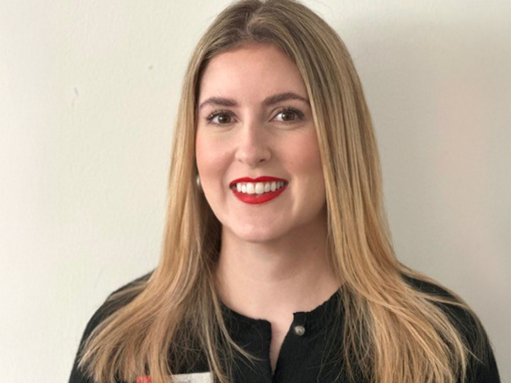 Femme aux longs cheveux blonds et au rouge à lèvres rouge, portant un haut noir, souriant devant un fond clair et uni.
