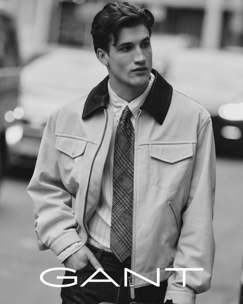 Un homme portant une chemise à col, une cravate et une veste claire marche à l'extérieur, les mains dans les poches. L'image est en noir et blanc, avec l'inscription "GANT" en bas.