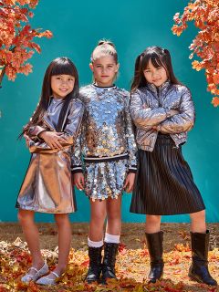 Trois jeunes filles se tiennent avec assurance au milieu des feuilles d'automne, vêtues de tenues brillantes et métalliques. Deux filles portent des vestes argentées, et la fille du milieu porte un haut et une jupe ornés de paillettes. Un fond bleu-vert contraste avec le feuillage d'automne orange.