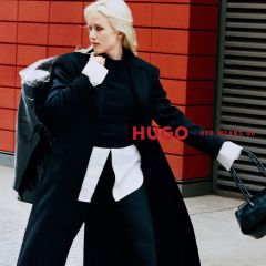 Une femme en manteau noir et chemise blanche pose à côté du lettrage « HUGO RED MEANS GO ».