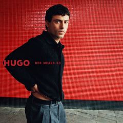 Un homme en cardigan noir et pantalon gris se tient devant un mur de carreaux rouges sur lequel est écrit « HUGO RED MEANS GO ».