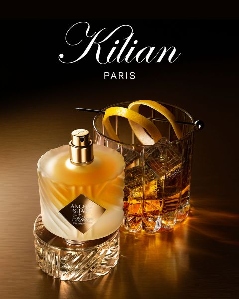Un flacon du parfum Kilian Paris Angels' Share est posé à côté d'un verre de whisky avec des glaçons et une garniture, le tout sur une surface brune réfléchissante avec un éclairage doux. Le logo Kilian Paris est visible en haut.