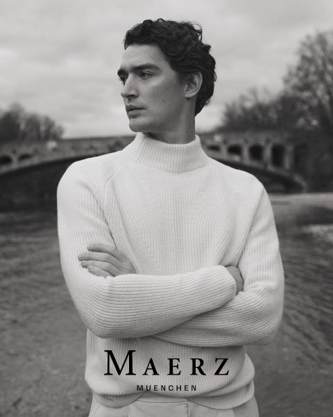 Un homme en pull en tricot blanc se tient les bras croisés au bord d'une rivière, avec un pont de pierre arqué et des arbres dénudés en arrière-plan. Le texte "MAERZ MUENCHEN" apparaît en bas de l'image en noir et blanc.