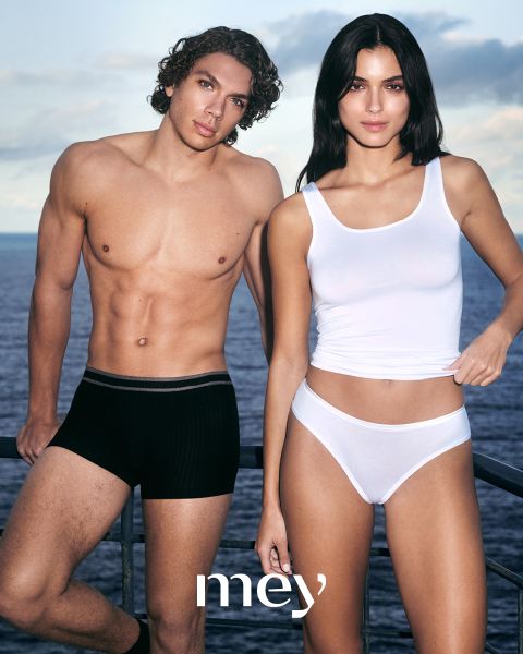 Un homme en caleçon boxer noir et une femme en débardeur blanc et sous-vêtements se tiennent côte à côte à l'extérieur, appuyés contre une balustrade, avec la mer et le ciel nuageux en arrière-plan. Le mot "mey" apparaît en bas.